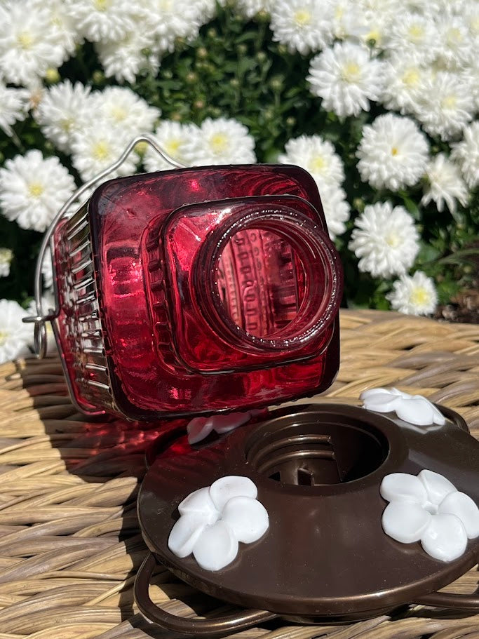 Red glass Hummingbird Feeder with decorative elements on a woven surface with flowers in the background