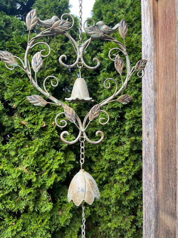 Decorative metal wind chime with floral designs and hanging bells against a green foliage background.