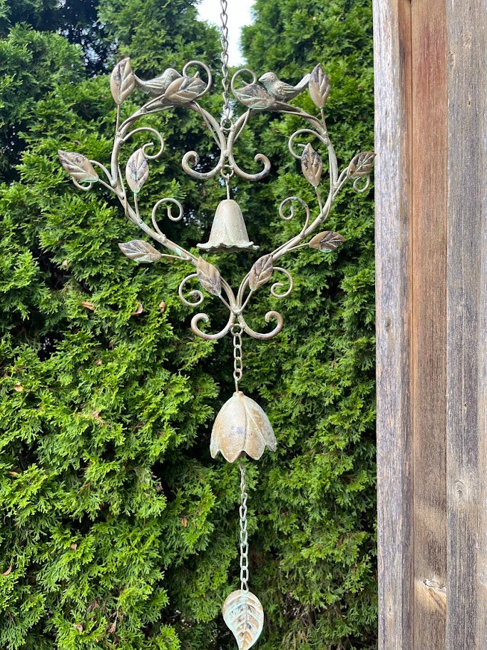 Decorative metal wind chime with leaves and a bell against a green bush background
