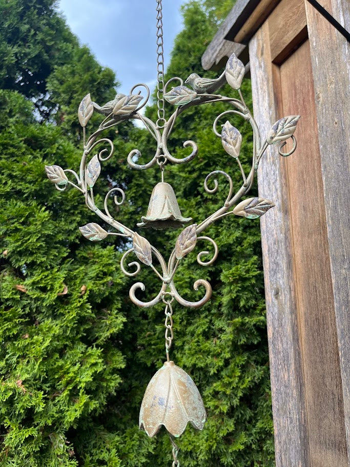 Decorative metal wind chime with leaf designs and a bell against a green foliage background.