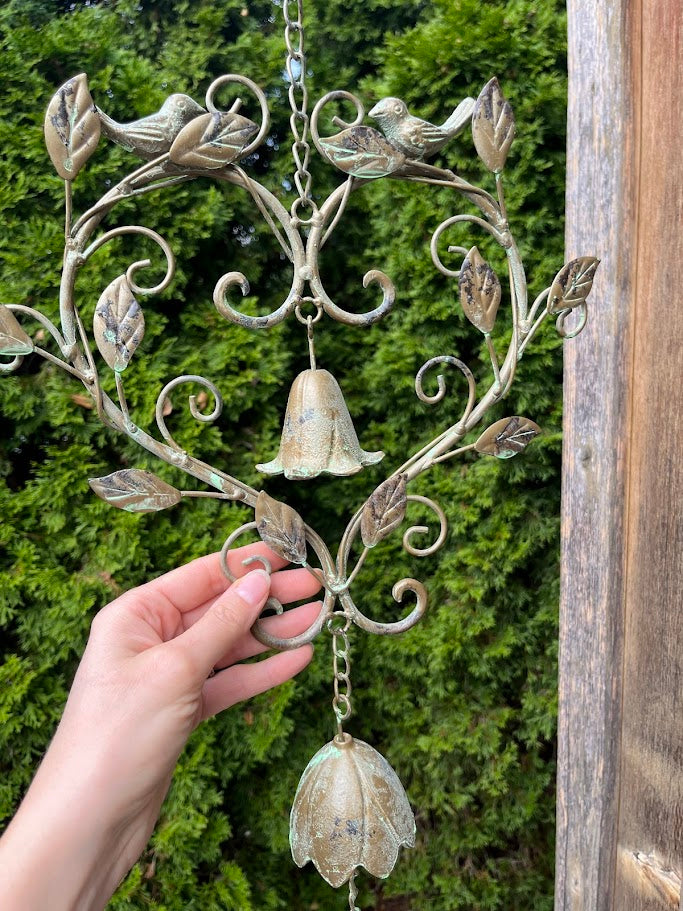 Decorative metal wind chime with floral designs and a hand holding it against a green background.