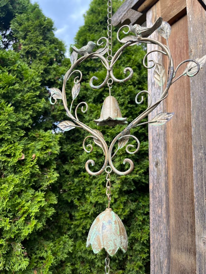 Decorative wind chime with floral design and hanging bells against a wooden post and green foliage background.