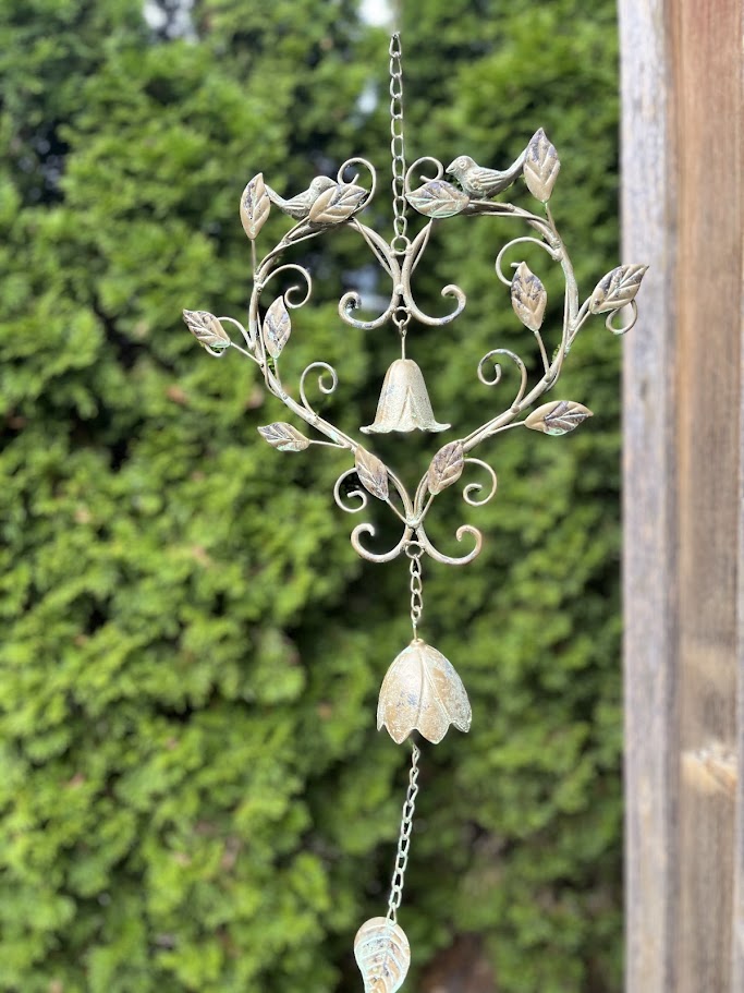 Decorative metal wind chime with floral designs against a green bush background