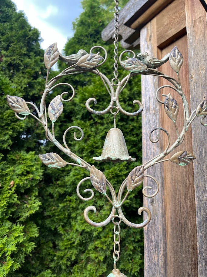 Decorative metal wind chime with leaf designs against a wooden post and green foliage background