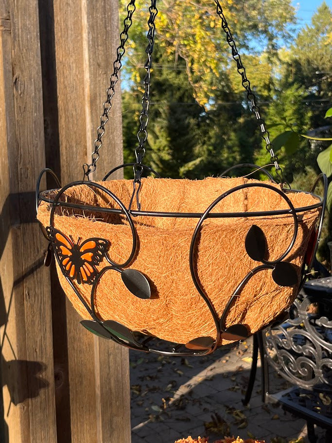 Decorative hanging planter with metal butterfly design against a wooden fence and garden background