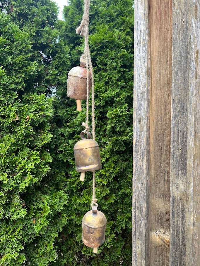 Three rustic metal bells hanging on a rope against a wooden post with green foliage in the background.