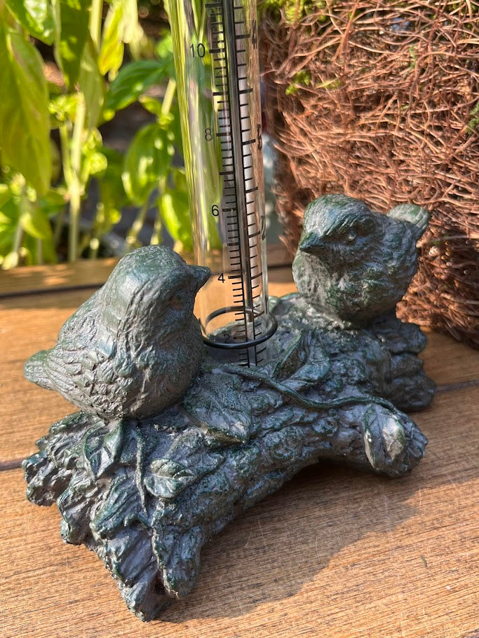 Decorative rain gauge with bird design on a branch, outdoors.