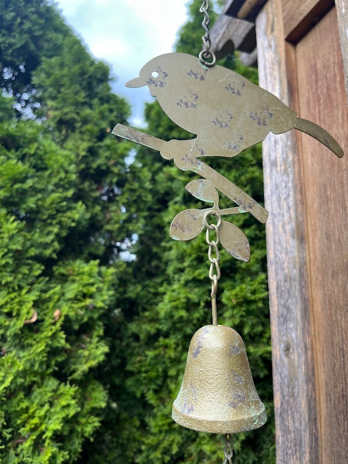 Decorative metal wind chime with bird design hanging on a wooden post against a green tree background.