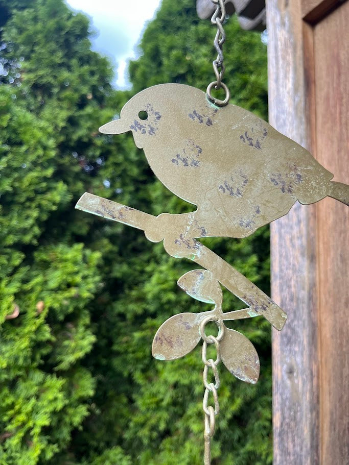 Decorative metal bird on a branch hanging against a wooden post with greenery in the background