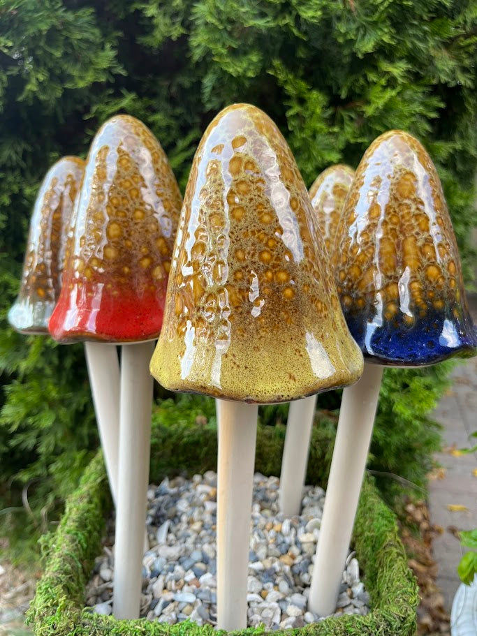 Set of ceramic mushroom decorations in various colors on a bed of pebbles with greenery in the background.
