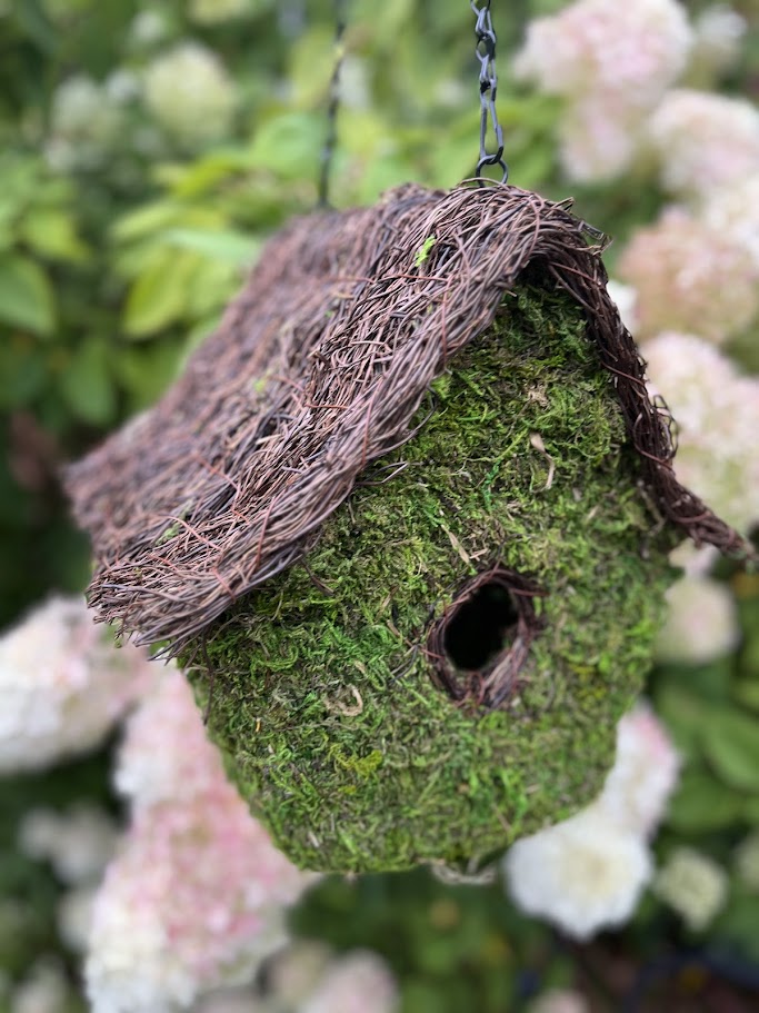 Moss-covered birdhouse with a thatched roof hanging among white flowers.