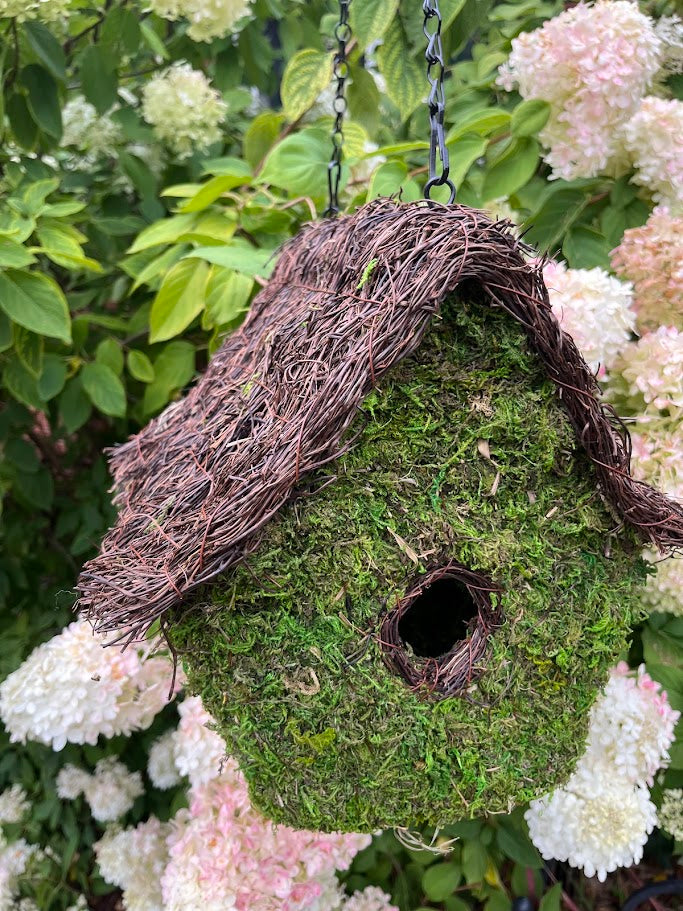 Moss-covered birdhouse with a natural twig design, surrounded by white flowers and green foliage.