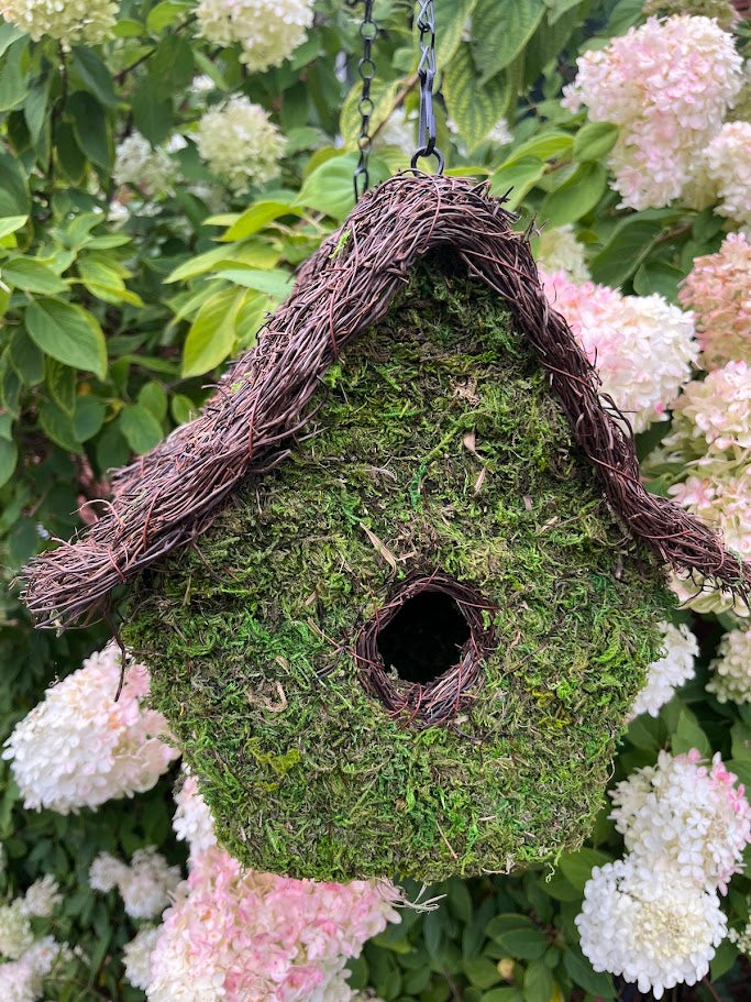 Moss-covered birdhouse with a natural twig roof, surrounded by white flowers and green leaves.