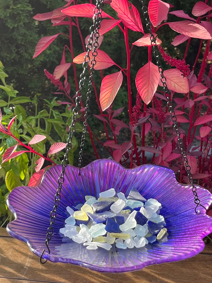 Purple glass bird bath with pebbles surrounded by pink and red foliage