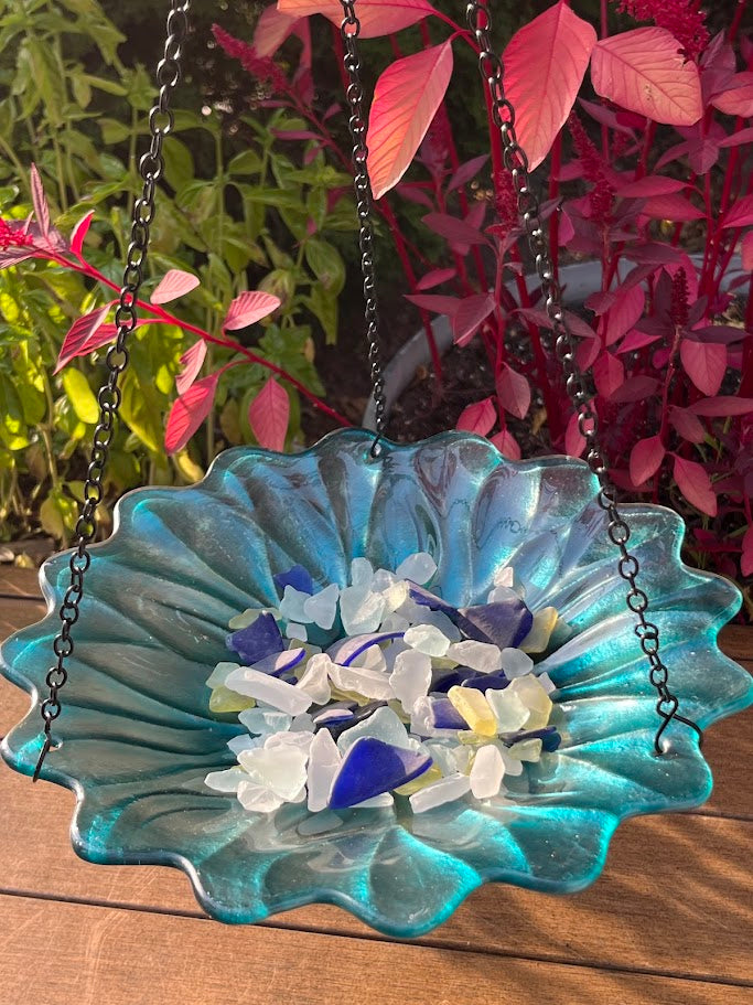 Blue glass bird bath with pebbles on a wooden surface with plants in the background
