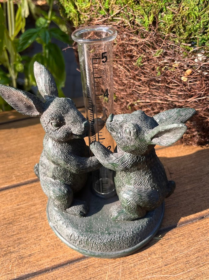 Decorative garden statue of two rabbits with a rain gauge, placed on a wooden surface.
