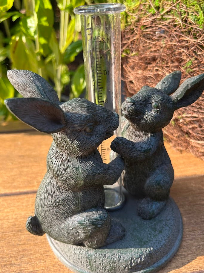 Decorative garden statue of two rabbits with a rain gauge on a wooden surface.