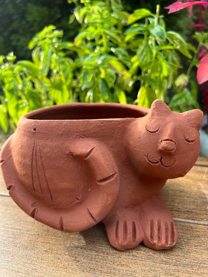 Terracotta cat-shaped planter on a wooden surface with green foliage in the background