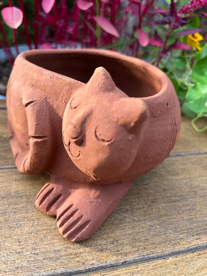 Terracotta pot shaped like a cat on a wooden surface with plants in the background