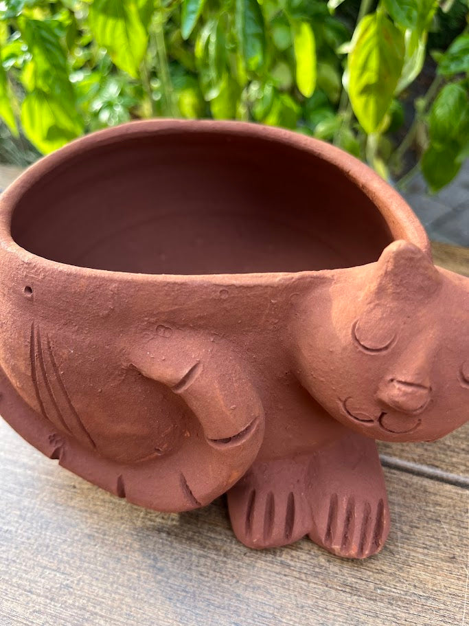 Terracotta pot shaped like a cat on a wooden surface with greenery in the background