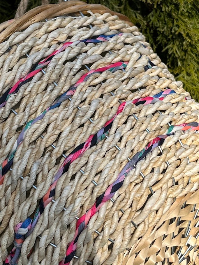 Close-up of a spool of multicolored ribbon on a woven surface with greenery in the background.