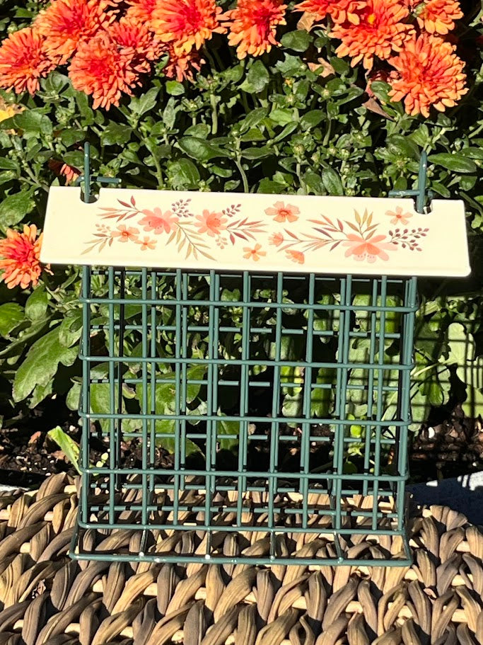 Decorative bird feeder with floral design on a green wire mesh background, surrounded by orange flowers.