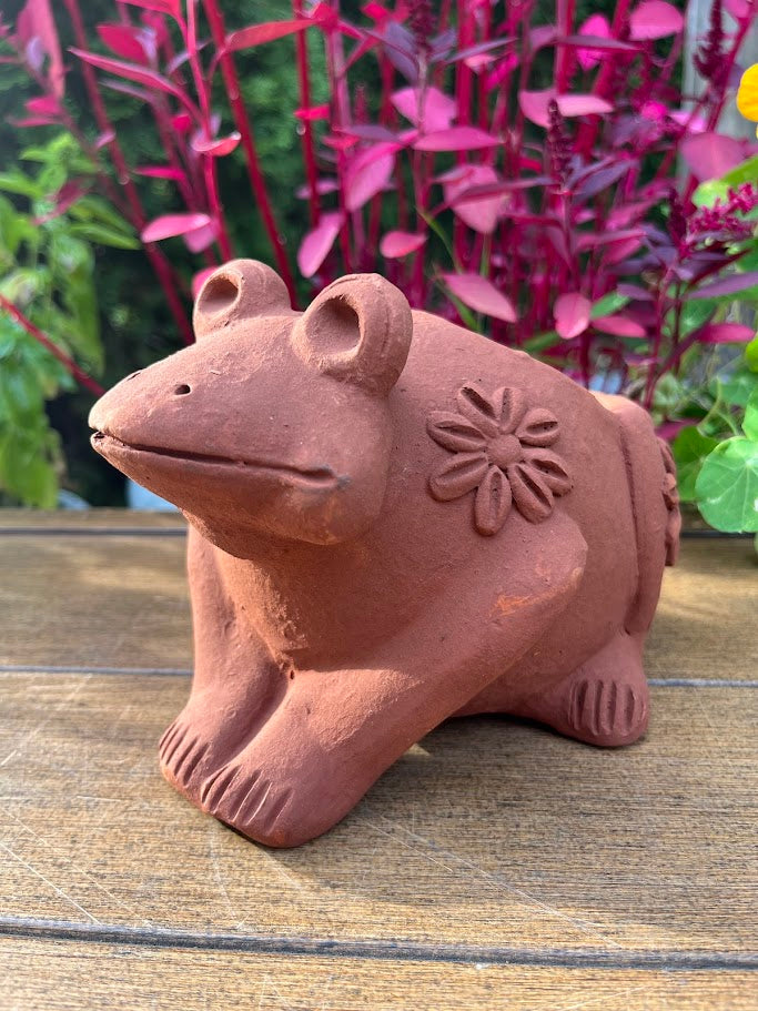 Terracotta frog figurine with floral design on a wooden surface with plants in the background