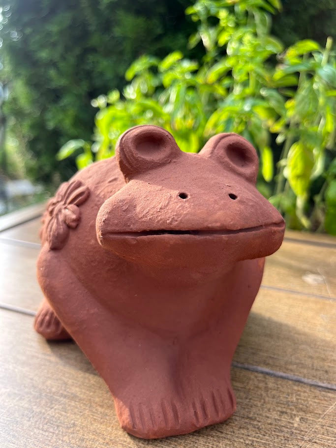 Terracotta frog figurine on a wooden surface with greenery in the background