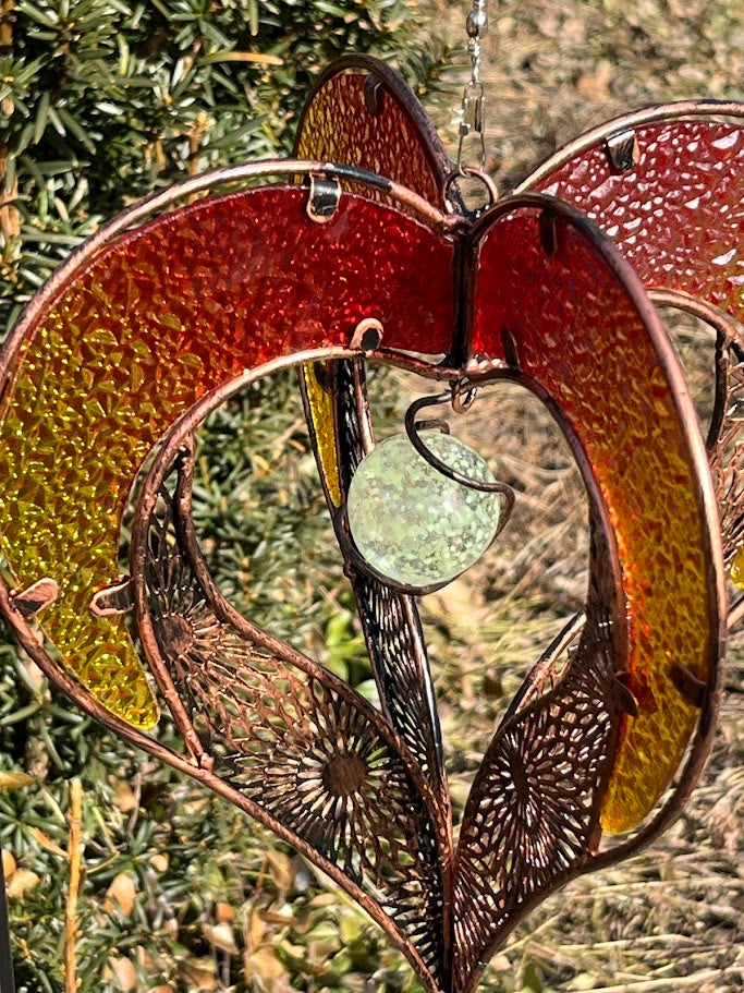 Decorative glass heart with floral patterns and a central round piece, hanging against a natural background.