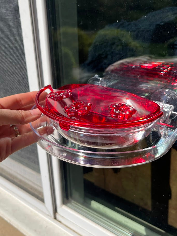 Red hummingbird feeder attached to a glass window with hand opening the lid