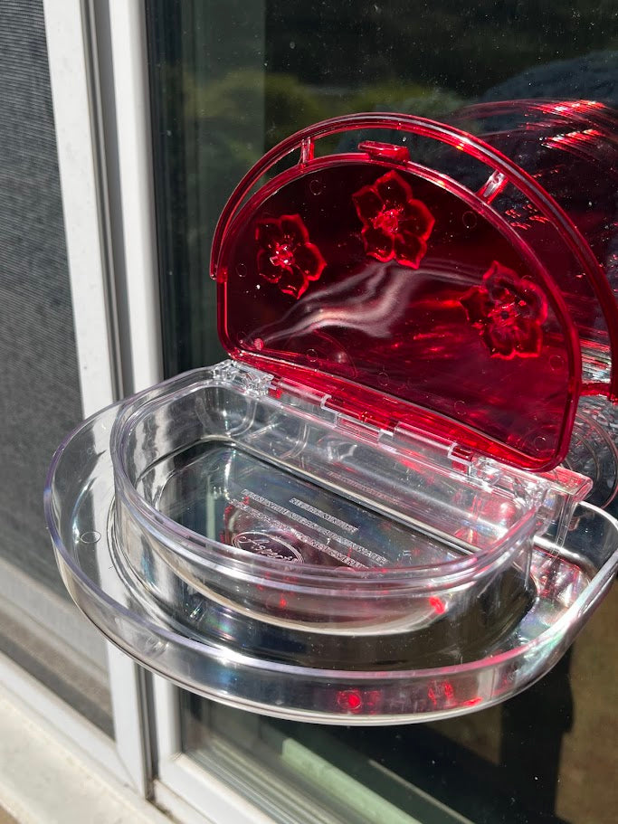 Red hummingbird feeder attached to a glass window with lid opened