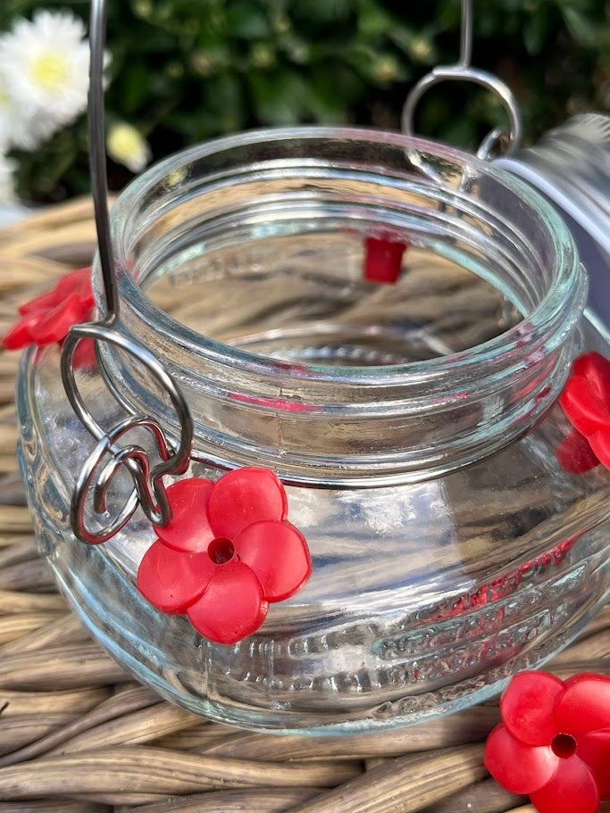 Glass mason jar hummingbird feeder on a woven surface with white flowers in the background