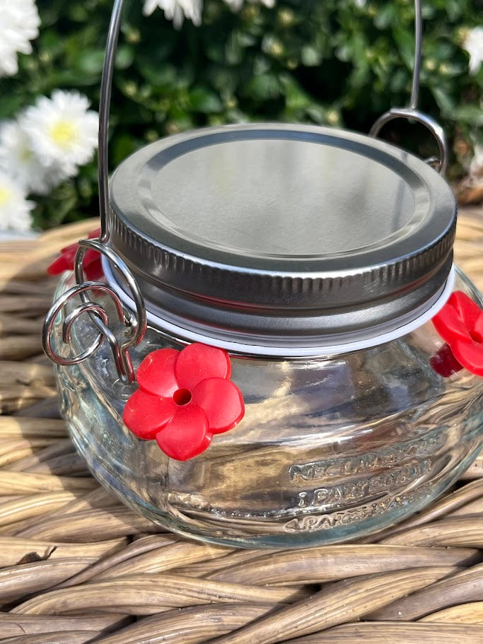 Glass mason jar hummingbird feeder on a woven surface with white flowers in the background