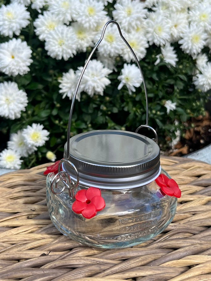 Glass mason jar hummingbird feeder on a woven surface with white flowers in the background