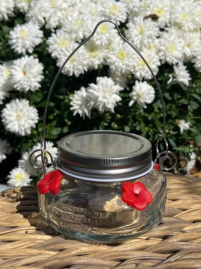 Glass mason jar hummingbird feeder on a woven surface with white flowers in the background