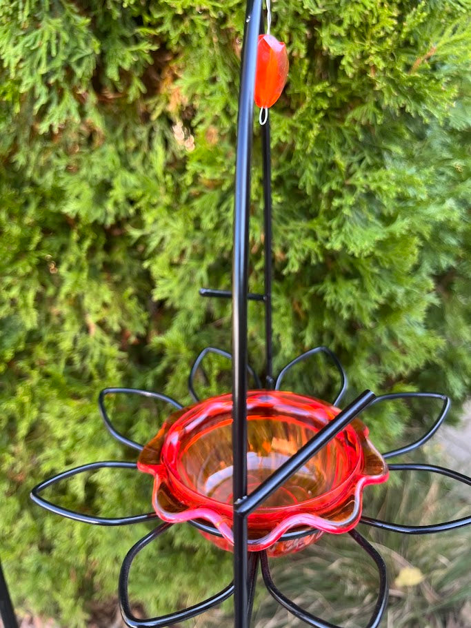 Red hummingbird feeder with black metal stand against a green bush background