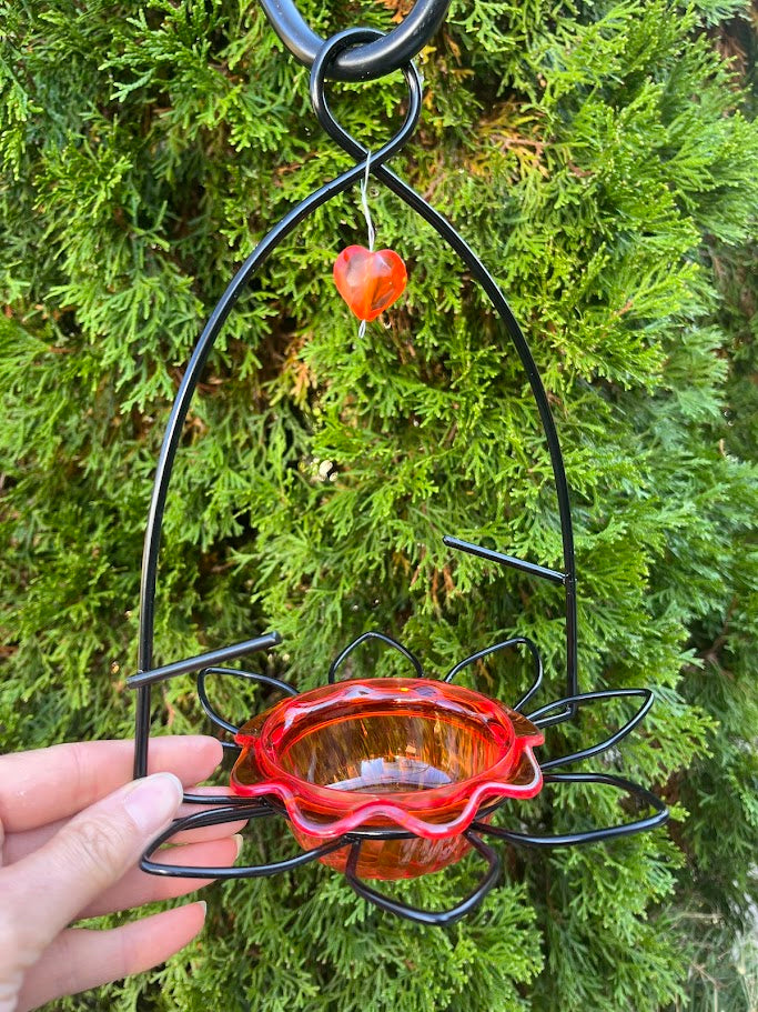 Hand holding a red bird feeder with black metal stand against a green bush background