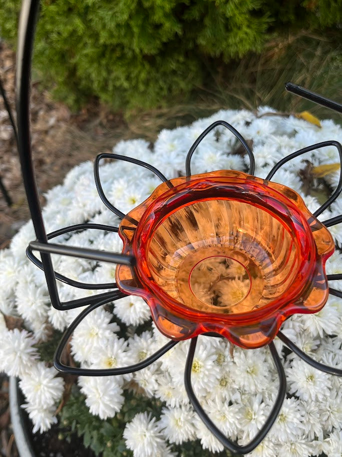 Amber glass hummingbird feeder with black metal stand against a white flower background