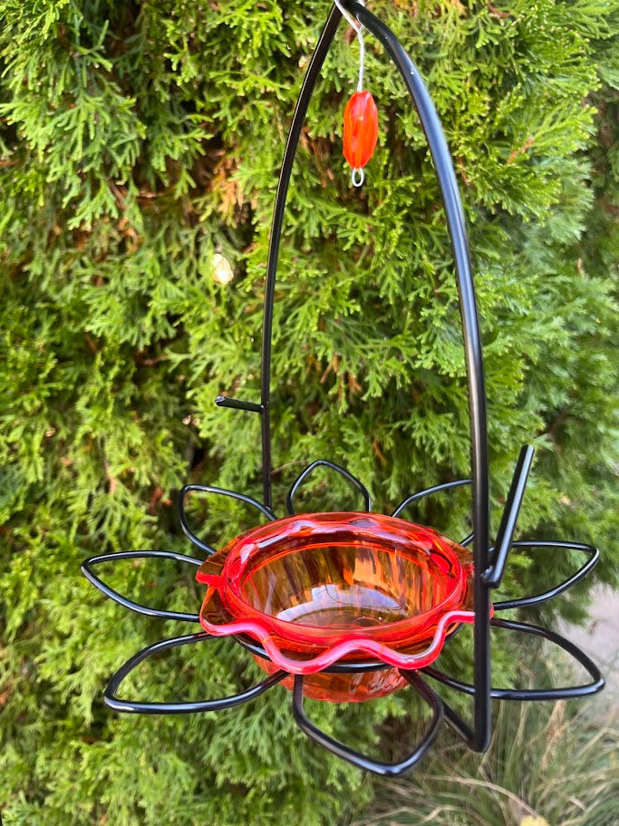 Hummingbird feeder with red dish and black metal stand against a green foliage background