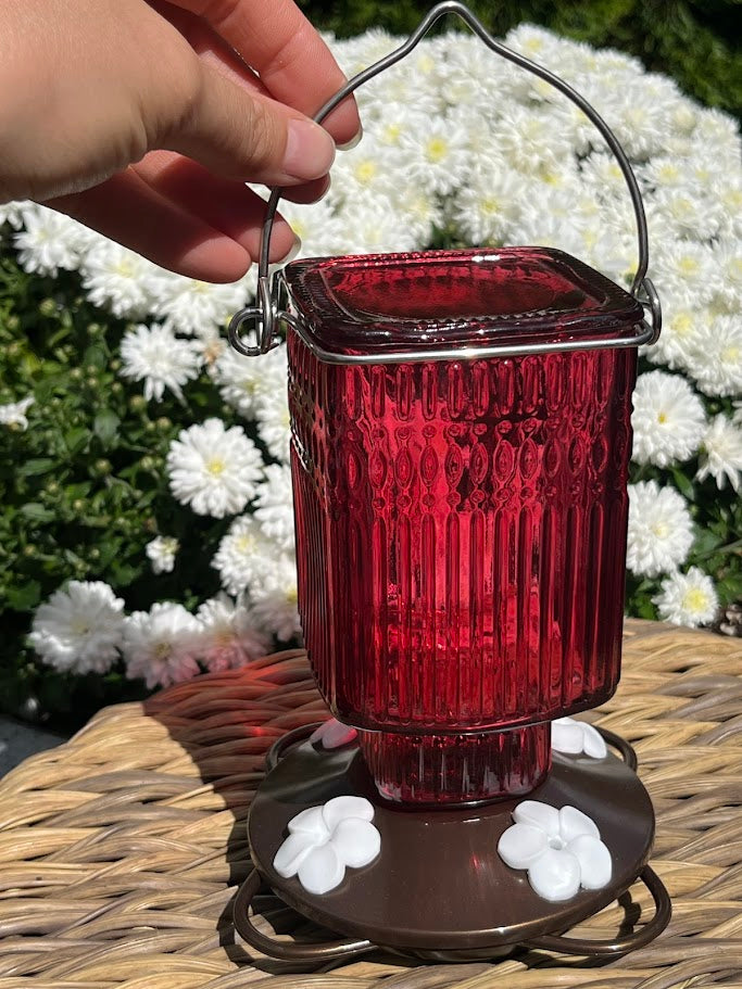 Red glass Hummingbird Feeder with decorative elements on a woven surface with flowers in the background