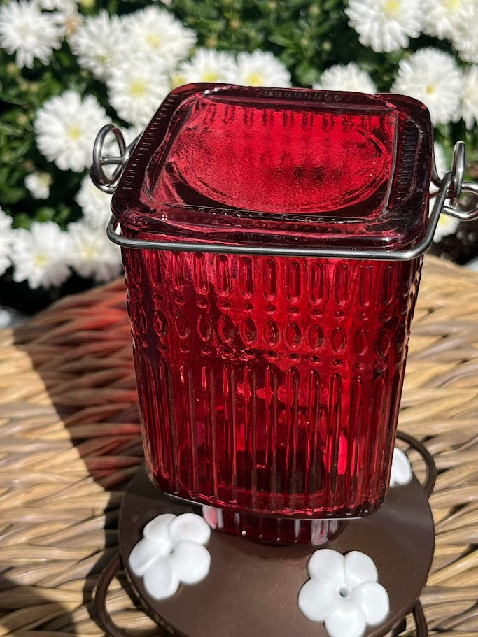 Red glass Hummingbird Feeder with decorative elements on a woven surface with flowers in the background