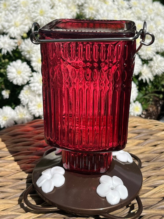 Red glass Hummingbird Feeder with decorative elements on a woven surface with flowers in the background