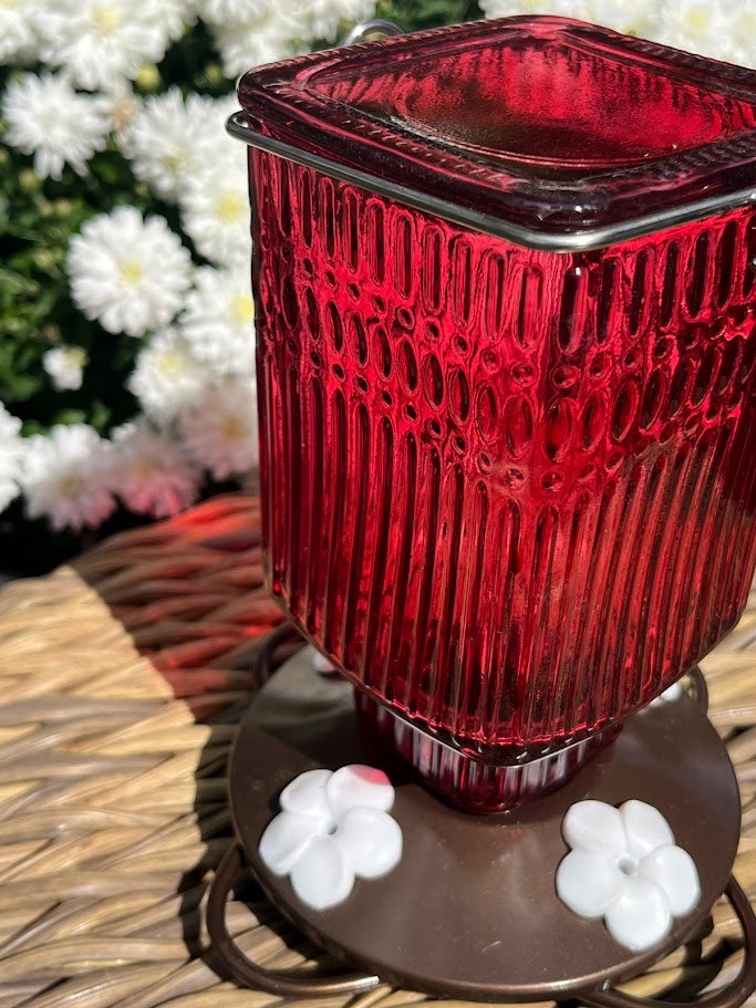 Red glass Hummingbird Feeder with decorative elements on a woven surface with flowers in the background