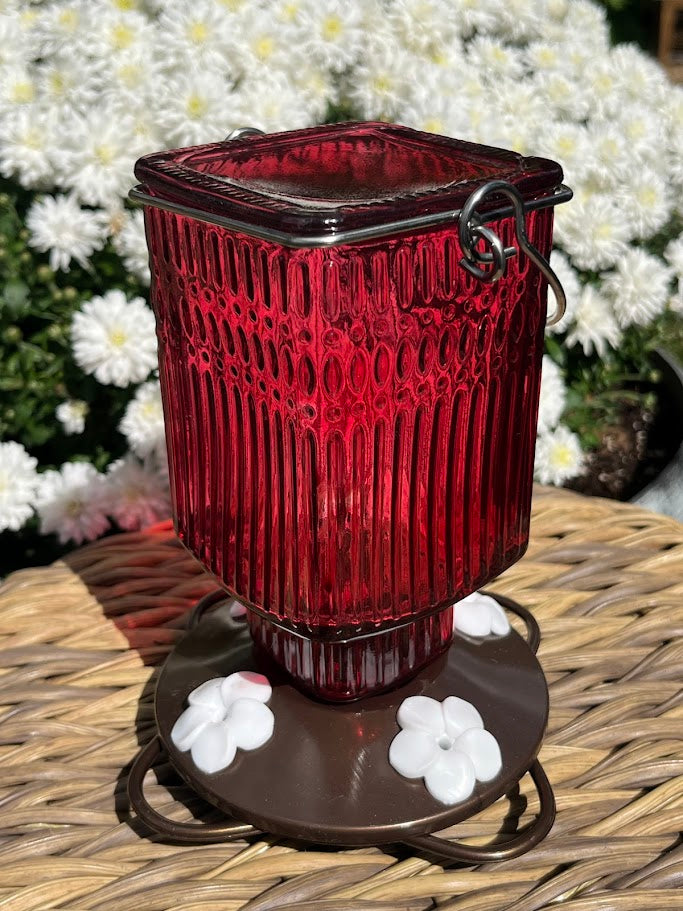 Red glass Hummingbird Feeder with decorative elements on a woven surface with flowers in the background