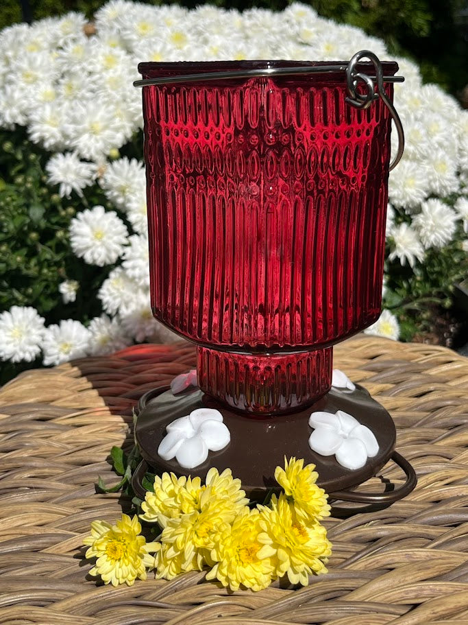 Red glass Hummingbird Feeder with decorative elements on a woven surface with flowers in the background