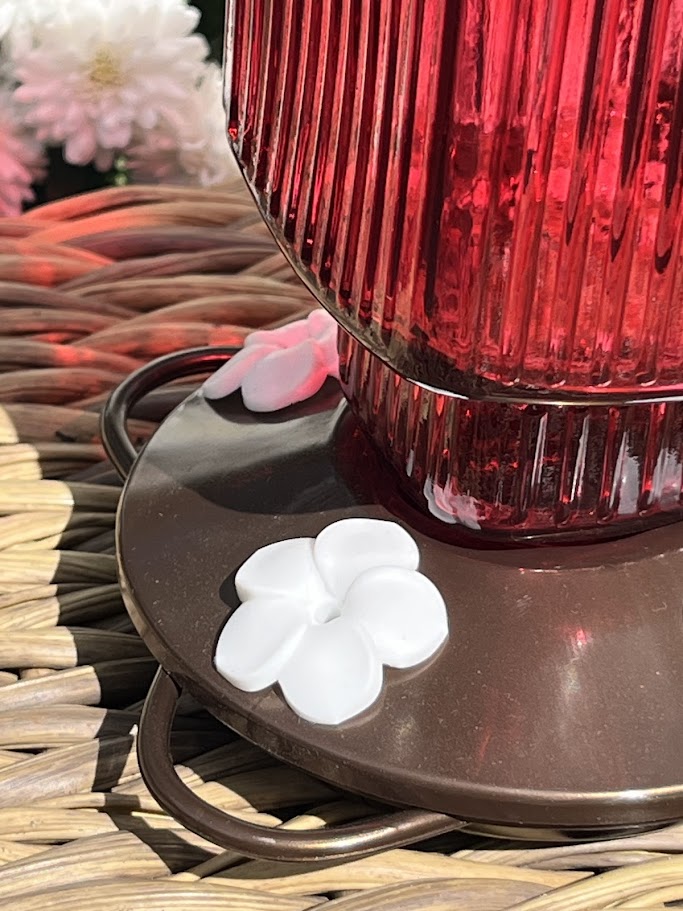 Red glass Hummingbird Feeder with decorative elements on a woven surface with flowers in the background