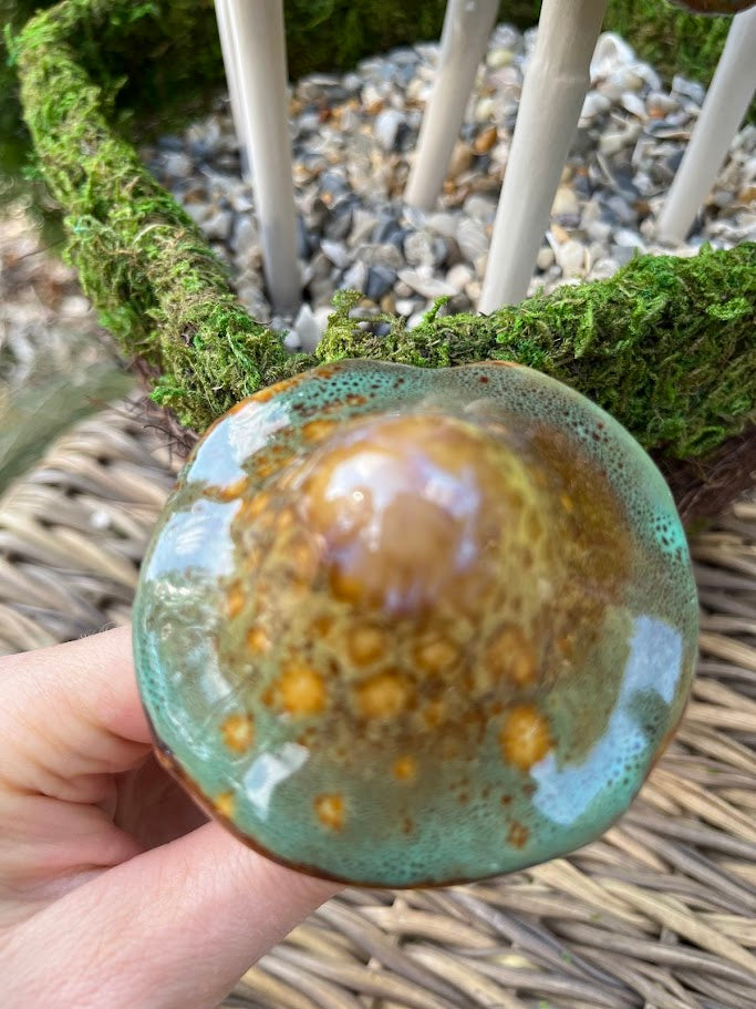 Hand holding a ceramic disc with green and brown glaze against a natural background