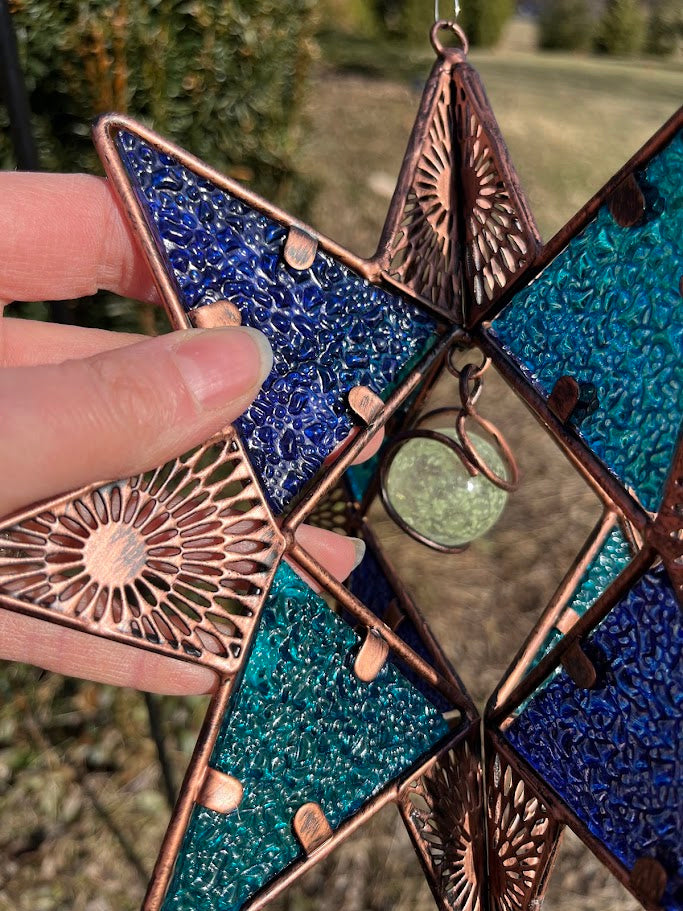Decorative star-shaped ornament with blue and green beads and copper accents held by a hand.