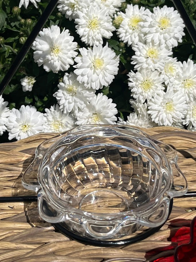 Clear glass hummingbird feeder bowl on a woven surface with white flowers in the background