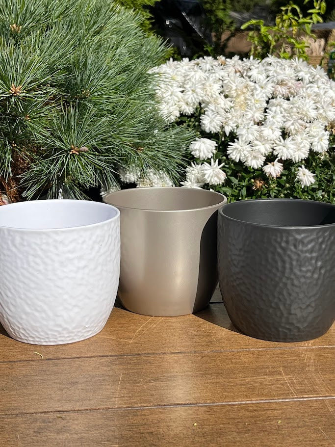 Three decorative pots in white, beige, and gray on a wooden surface with plants in the background.